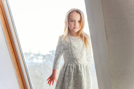 小可爱的可爱的微笑的女孩, 白色的礼服站在窗台上, 在明亮的客厅在家里思考