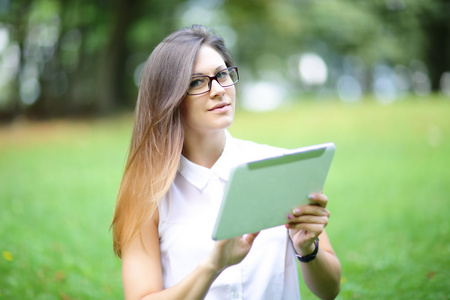 年轻的女人，使用平板电脑和手机在绿色的田野，公园与阳光的女孩