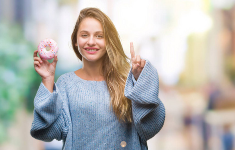 年轻美丽的金发女子吃甜甜甜圈在孤立的背景惊讶与一个想法或问题指向手指与幸福的脸, 第一