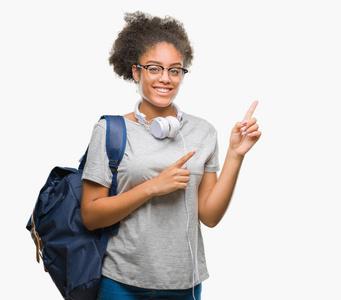 年轻的美国黑人学生妇女戴着耳机和背包在孤立的背景下微笑着, 看着两只手和手指指向侧面的相机