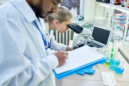 看显微镜的年轻女性科学家的侧面画像在实验室进行医学研究时, 同事在前台对剪贴板做笔记