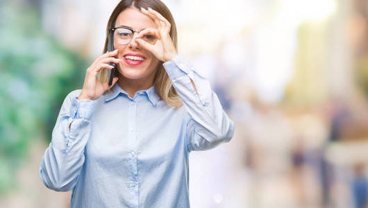 年轻美丽的女商人说话打电话使用智能手机在孤立的背景与快乐的脸微笑做确定的标志与眼睛看着手指