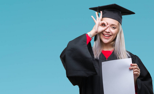 年轻的金发女子穿毕业制服持有学位, 在孤立的背景和快乐的脸微笑做 ok 标志与手在眼睛看通过手指