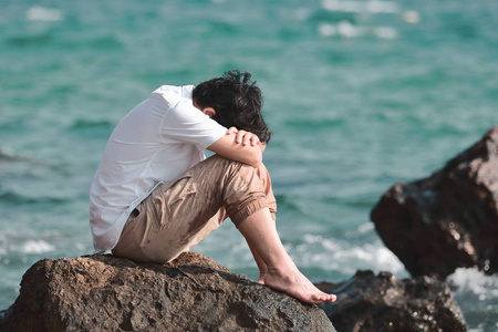 忧郁的年轻亚裔男子抱着膝盖坐在海边的岩石上