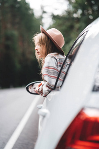 英俊的女人坐在车上看着窗外的美丽景色在森林里。漂亮的女孩喜欢乡村路的时髦。生活方式概念