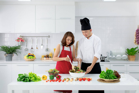 美丽的亚洲妇女在红色围裙学习如何烹饪和混合蔬菜沙拉在大碗, 由亚洲厨师在白色的现代厨房教学