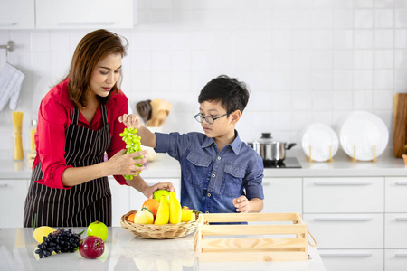 美丽的亚洲妇女在红色衬衫和黑色围裙教她的儿子如何安排假水果和蔬菜装饰在白色干净的厨房