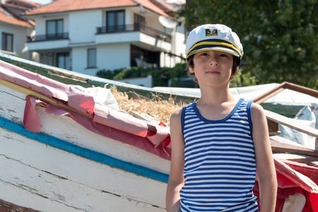 一个男孩的肖像在条纹 t恤衫和船长的帽子上的背景上的一条老木船
