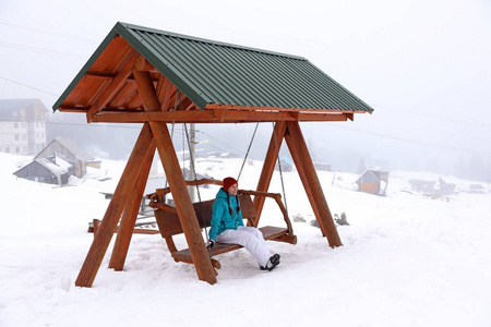 美丽的女人坐在雪地度假胜地的摇摆长椅上。寒假