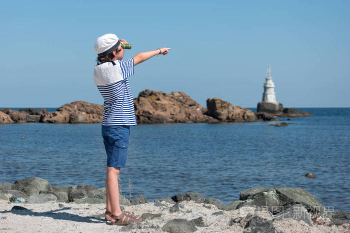 穿条纹 t恤衫的男孩和海滩上的帽子, 看着望远镜, 向大海展示他的手