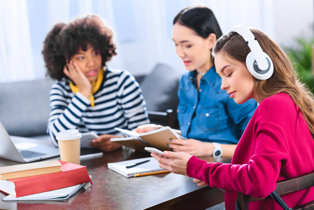 年轻学生在耳机中的选择性焦点使用智能手机与多民族朋友附近
