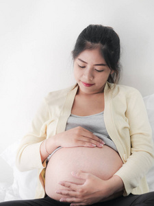 年轻的亚洲怀孕妇女抚摸她的腹部, 坐在床上, 生活方式的概念