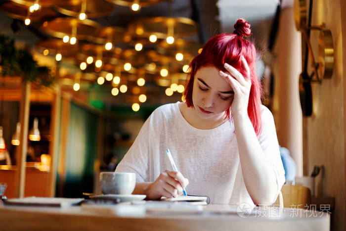 集中的年轻妇女在笔记本上写, 而坐在餐桌上咖啡馆.