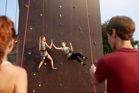 微笑的女孩给了高五挂绳在人工攀登训练墙上, 并由朋友在保护束保险。攀岩安全系统。微笑的妇女在抱石赛练习
