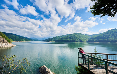 摄影师女性在湖 Vidraru 喀尔巴阡山罗马尼亚。阳光明媚的一天美丽的风景 高山, 森林, 湖泊, 蓝天白云