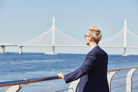 眼镜和深色夹克上的金发男人的背影欣赏夏季水面倾斜金属扶手, 在背景