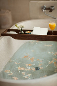 明亮的浴室在家里。用花瓣和盐沐浴。托盘在浴缸与橙汁, 记事本和蜡烛