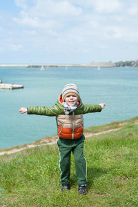 滑稽的小男孩穿着温暖的夹克围巾和帽子跳上绿色斜坡反对海港的背景与小船在寒冷的春天