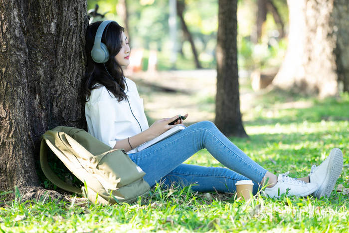 年轻美丽的亚洲妇女放松和听音乐使用耳机在草地上的公园生活方式和