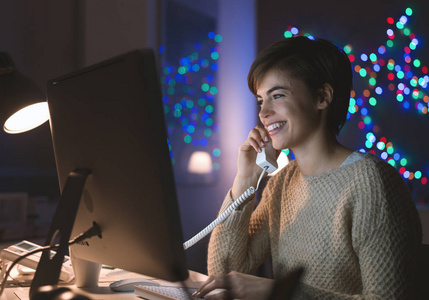 年轻的微笑的妇女工作与她的电脑和电话交谈深夜, 她拿着听筒