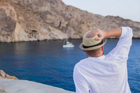 一个戴着帽子和白衬衣的男人坐在海边, 凝视着游艇.照片