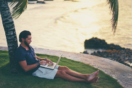 在棕榈树下热带海滩上的笔记本电脑工作的年轻人