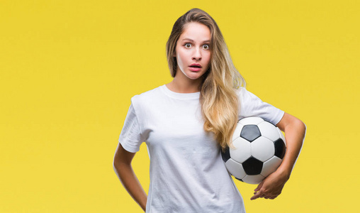 年轻美丽的金发女子持有足球在孤立的背景惊吓与惊讶的脸, 害怕和兴奋与恐惧表达