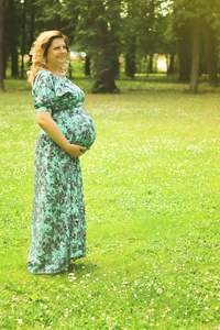 一个年轻漂亮的孕妇抱着她的手在她的胃在一个长裙的背景下的树木和绿色肝在夏季