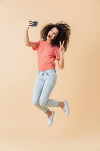 满长的肖像一个开朗的年轻女孩采取自拍的手机, 而跳跃隔离在米色背景