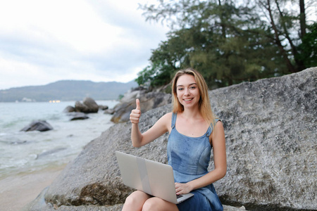 年轻妇女使用膝上型电脑和坐在石头附近的海, 空的早晨海滩, 显示竖起大拇指