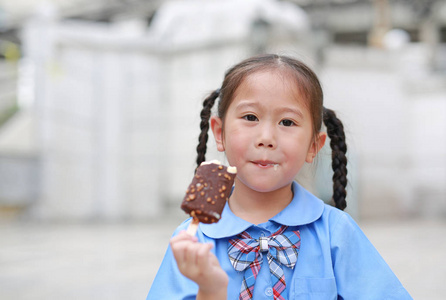 愉快的小亚洲儿童女孩在学校制服享受吃巧克力香草冰淇淋户外