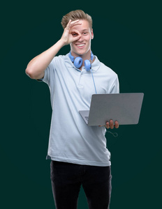 年轻英俊的金发男子使用笔记本电脑与快乐的脸微笑做 ok 签署与手在眼睛看通过手指