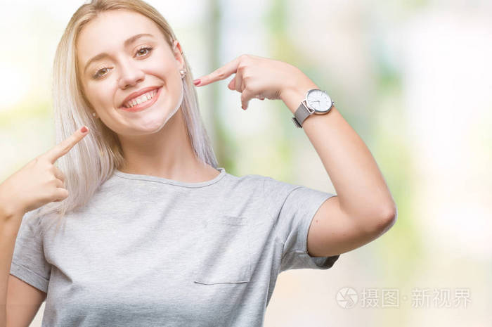 年轻的金发女人在孤立的背景下微笑着自信地显示和指着手指的牙齿和