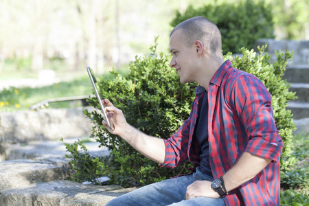 英俊的年轻人坐在公园里, 用平板电脑享受户外活动。选择性对焦和小景深