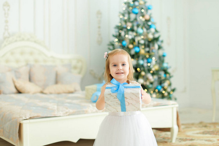 小女孩保持礼物, 穿白色礼服和站立在卧室用圣诞树