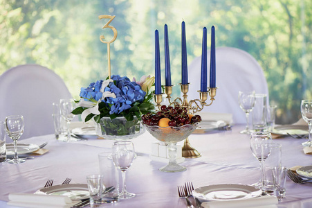 表与经典设计。在一个高花瓶与绣球和百合的组合, 一个金色烛台与蓝色蜡烛。装饰和植物。碗樱桃