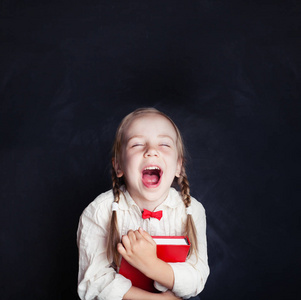快乐的小女孩笑了。快乐的孩子与红色书在空白黑板背景与拷贝空间。返回学校概念