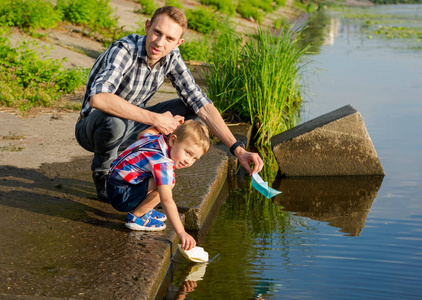 爸爸帮助小男孩把纸船放到水里。爸爸和一个小儿子夏天在河里发射纸船