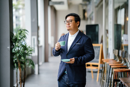 一位受过教育的中年中国亚裔男子在商务休闲和眼镜的肖像。在城里的白天, 他拿着一个优雅的杯子和碟子喝茶。