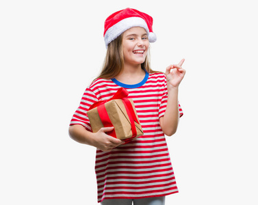 年轻美丽的女孩戴着圣诞帽, 拿着礼物在孤立的背景非常高兴用手和手指指向一边
