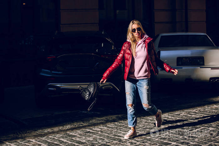 街头时尚概念一个美丽的女孩的肖像坐在外面, 蓝色牛仔裤, 太阳眼镜, 硬灯, 红色夹克, 秋季天气
