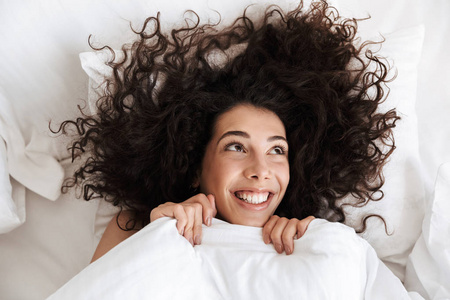 从上面的欢快的女人的肖像20s 与深色卷曲的头发看着向上, 而躺在床上的白色亚麻与快乐的笑容