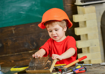 可爱的孩子玩工作工具。戴橙色防护头盔的男孩拿着一块木块。小建设者锤钉