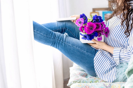 快乐的女人在家中带着一束鲜花和一份礼物, 在沙发上靠近窗户。盛开的花朵。卡。文本的可用空间