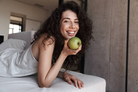 华丽的卷发妇女20s 与长的深色头发穿着丝绸休闲服装吃新鲜的绿色苹果与乐趣和微笑, 而躺在沙发上在家