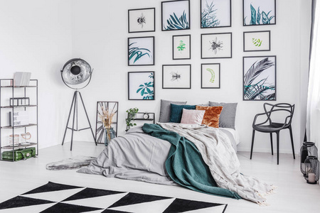 一张床的真实照片与垫子和毯子站在一个单色卧室内部的椅子, 灯和架子, 旁边的墙上有植物学海报