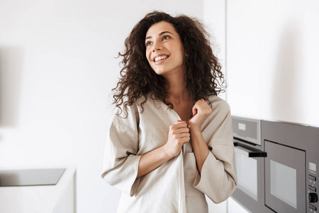 黑头发的白种卷发妇女的相片穿着丝绸休闲服装微笑着看一边站在厨房附近的烹饪场所
