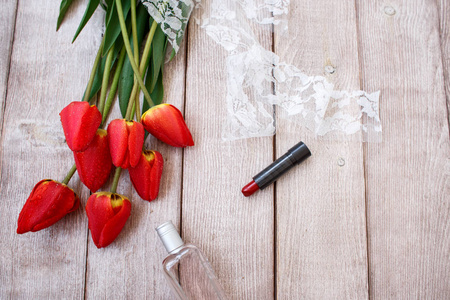 红色郁金香洒在木质背景和红色口红和一瓶厕水上。女装配件。一束新鲜的红色郁金香系着白色花边丝带