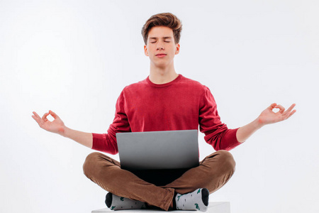 年轻的男人学生闭着眼睛坐在莲花的位置与笔记本电脑在他的腿在白色背景