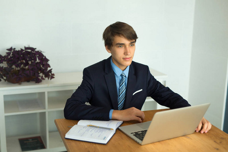 漂亮的年轻的家伙商人在西装坐在桌子上的办公室与笔记本电脑